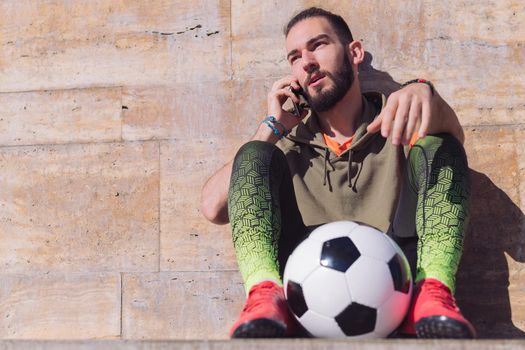 handsome sportsman talking on the phone while resting next to his soccer ball, concept of technology and urban sport lifestyle in the city, copy space for text