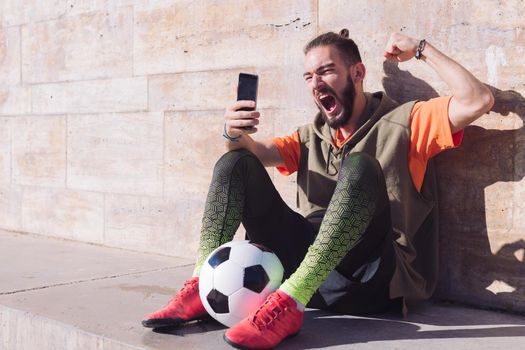 hipster football fan celebrates a victory when he is checking his smart phone, concept of technology and urban sport lifestyle in the city, copy space for text