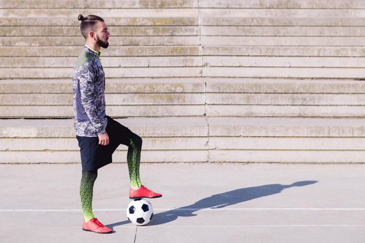 handsome sportsman in a concrete soccer court with hair in a bun and a foot on his football ball, concept of healthy lifestyle and urban sport in the city, copy space for text