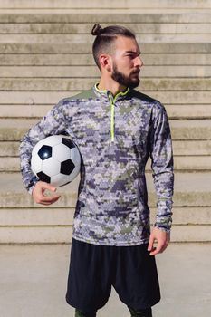 vertical photo of an attractive hipster sportsman in a concrete soccer court with the football ball under his arm, concept of healthy lifestyle and urban sport in the city