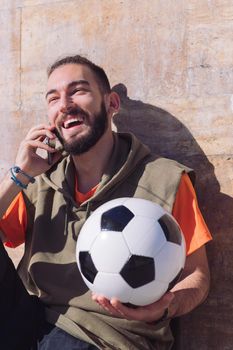 handsome sportsman laughing and talking by phone while resting with his soccer ball, concept of technology and urban sport lifestyle in the city, copy space for text