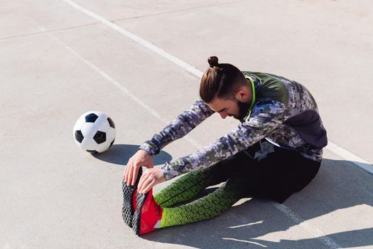 fashionable sportsman stretching legs sitting on the floor next to his football ball in a concrete soccer court, concept of healthy lifestyle and urban sport in the city, copy space for text