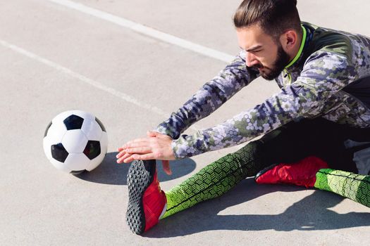 young footballer stretching legs sitting on the floor next to his football ball in a concrete soccer court, concept of healthy lifestyle and urban sport in the city, copy space for text