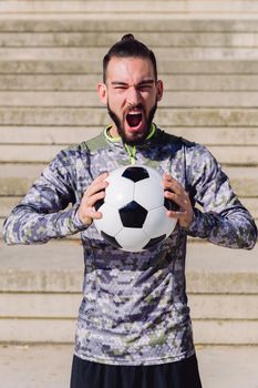 vertical portrait of a handsome sportsman screaming in a concrete soccer court with the football ball in his hands, concept of healthy lifestyle and urban sport in the city