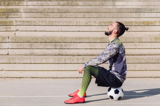 handsome sportsman with hair in a bun laughing in a concrete soccer court sitting on his football ball, concept of healthy lifestyle and urban sport in the city, copy space for text