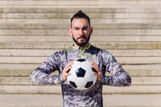 horizontal portrait of an attractive hipster sportsman in a concrete soccer court with the football ball in his hands, concept of healthy lifestyle and urban sport in the city, copy space for text