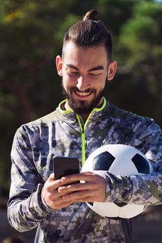 portrait of a happy sportsman with a soccer ball smiles writing a message on his mobile phone on the football court, concept of technology and urban sport lifestyle in the city