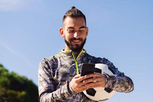 horizontal portrait of a happy sportsman with a soccer ball smiling writing a message on his mobile phone, concept of technology and urban sport lifestyle in the city, copy space for text