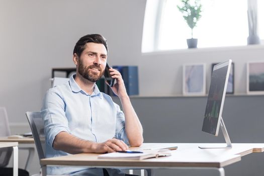 Young man with a beard, freelancer, manager, worker talking on a cell phone in the office, sitting at a computer desk, talking, waving