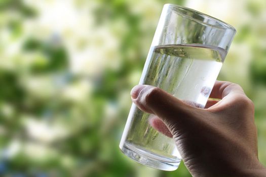 A glass of water in a hand close-up on a natural green background outdoors..