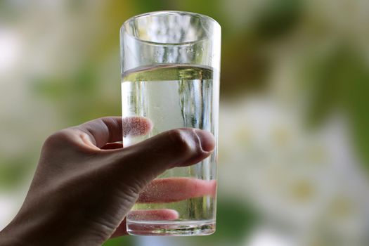 A glass of water in a hand close-up on a natural green background outdoors..