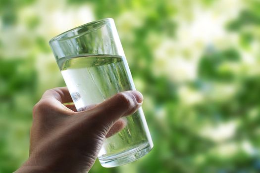 A glass of water in a hand close-up on a natural green background outdoors..