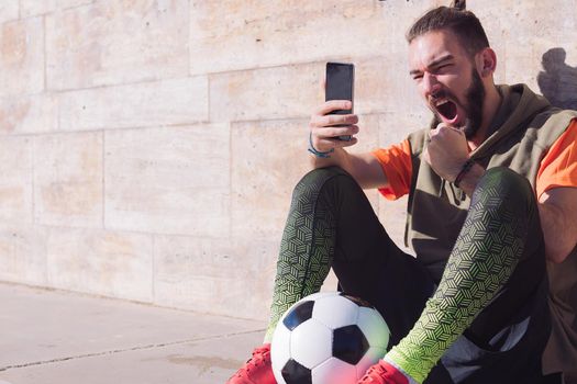 handsome football fan celebrates a victory when he is consulting his smart phone, concept of technology and urban sport lifestyle in the city, copy space for text