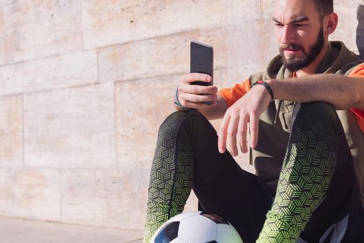 handsome football player rest sitting on the court consulting his smart phone, concept of technology and urban sport lifestyle in the city, copy space for text