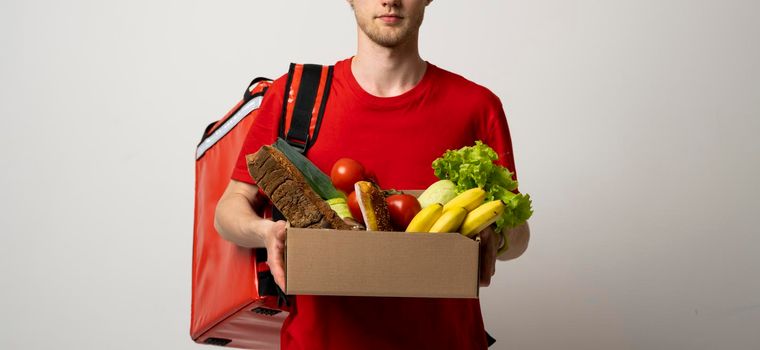 Crop picture of delivery man in red uniform carrying paper box with food products isolated over red background. Food delivery service