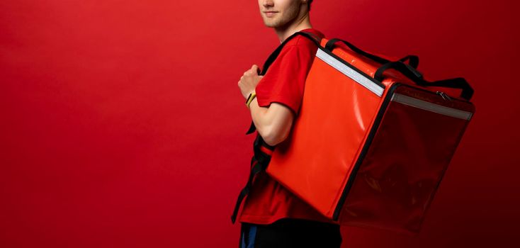 Portrait of young courier. Delivery man in uniform with thermo backpack isolated on red background. Fast home delivery. Online order