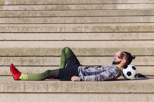 handsome sportsman with hair in a bun resting in a concrete soccer court with head on his football ball, concept of healthy lifestyle and urban sport in the city, copy space for text