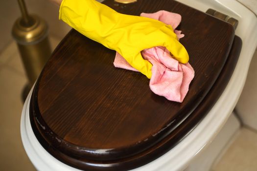 Female hands in yellow rubber gloves are holding a rag and wiping the toilet with detergent and disinfectant. The woman is cleaning the bathroom.