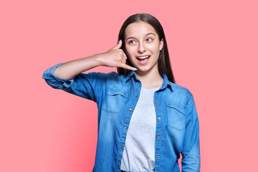 Smiling teenage female making phone gesture with hand and fingers, showing telephone receiver, sign symbol call me. Beautiful teen girl posing on red pink color studio background communication concept