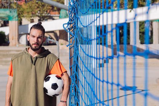 portrait of an attractive hipster football player by the goal net with the ball under his arm, concept of healthy lifestyle and urban sport in the city, copy space for text