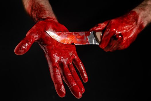 A man with blood-stained hands cuts his palm with a knife on a black background