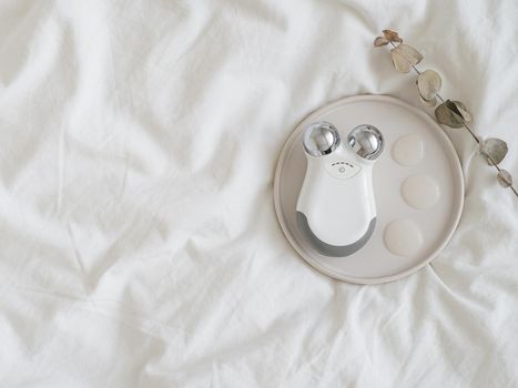 Microcurrent concept. Face massager with micro currents and gel primer drops. Home use device for anti-aging and face lifting on white bed. Top view or flat lay, copy space for text.