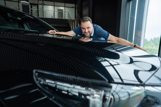 Happy caucasian man hugging the hood of his new car in a car dealership