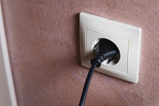 Electric socket on a pink wall. The black wire plug is connected. Renovated backdrop of studio apartment. Blank copy space single white plastic socket.