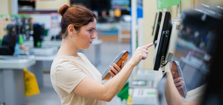 A frustrated woman uses a self-checkout counter. The girl does not understand how to independently buy groceries in the supermarket without a seller.