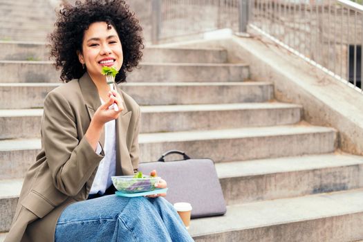 happy young woman eating a salad from a lunch box sitting on a staircase during a break from work, concept of urban lifestyle and healthy food, copy space for text