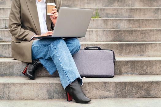 unrecognizable young woman works outdoors with her laptop computer sitting on a staircase while drinking coffee, concept of business and urban lifestyle, copy space for text