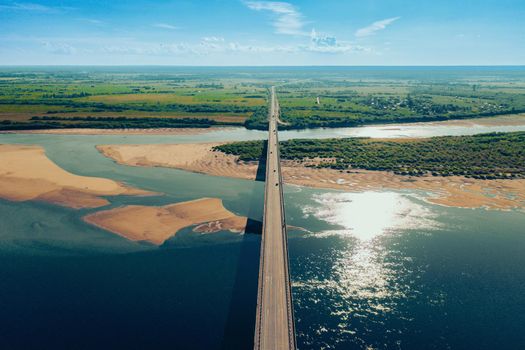 Aerial drone shot of cars driving on bridge over river and sandy riverside at sunset. Amazing aesthetic 4k top view shot with beautiful coast with dune sands. Drone aerial bridge with car traffic