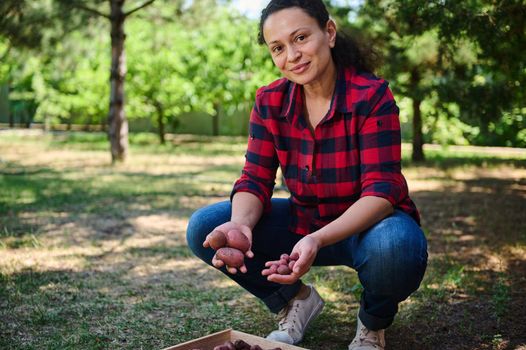 Portrait of a young successful multi-ethnic woman eco farmer, gardener, agronomist smiles looking at camera while posing with freshly harvested crop of organic potatoes, for sale in farmers markets