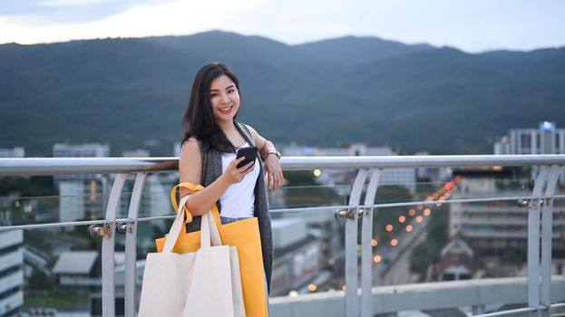 Pretty asian woman with shopping bags standing on rooftop terrace with cityscape at sunset.