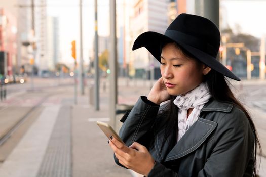portrait of an elegant young asian woman consulting cell phone while waiting at the tram stop, concept of public transport, technology and urban lifestyle