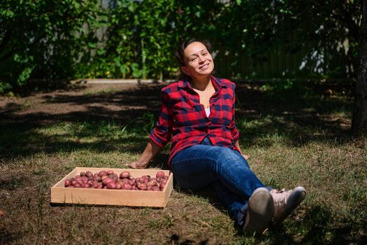 Pleasant multiethnic woman, farmer agronomist in a plaid shirt and blue denim jeans, sitting on the grass in the countryside house backyard, resting after harvesting crop of fresh organic potatoes