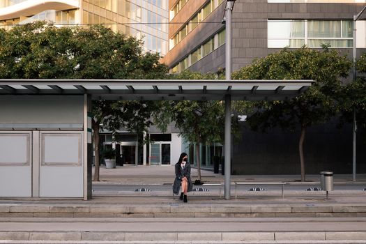 tram stop with a woman wearing a protective mask sitting and waiting for the train, concept of public transportation and urban lifestyle