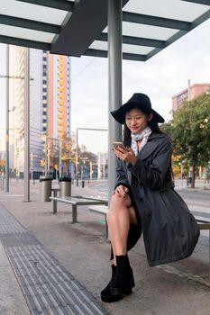 vertical photo of an elegant young asian woman consulting cell phone while waiting at the bus stop, concept of public transport, technology and urban lifestyle