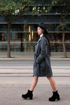vertical photo of a classy asian woman walking in the city with hat and trench coat, concept of elegance and urban lifestyle, copy space for text