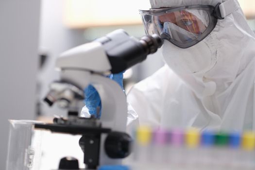 Biologist in protective suit with goggles and medical mask working with microscope in laboratory. Research dangerous biological viruses concept