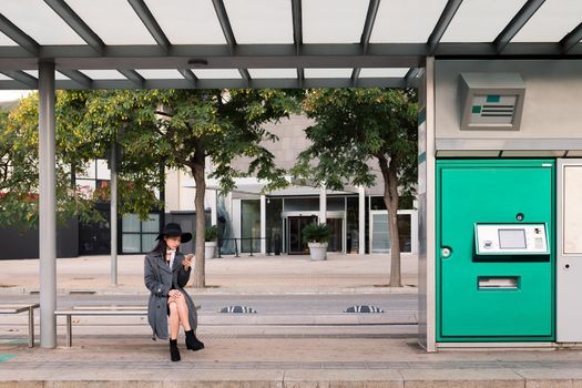 elegant asian woman consulting the phone while waiting at the bus stop, concept of public transport, technology and urban lifestyle