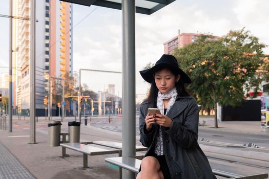 elegant young asian woman looking at her cell phone while waiting at the bus stop, concept of public transport, technology and urban lifestyle