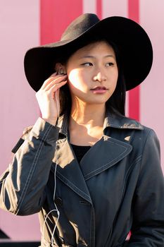 vertical portrait of a young beautiful classy asian woman with hat putting on earphones in a pink background, concept of technology and fashion lifestyle