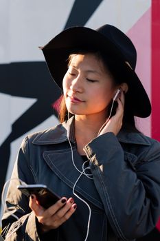 vertical portrait of a young beautiful classy asian woman with earphones listening music from her phone, concept of technology and fashion lifestyle
