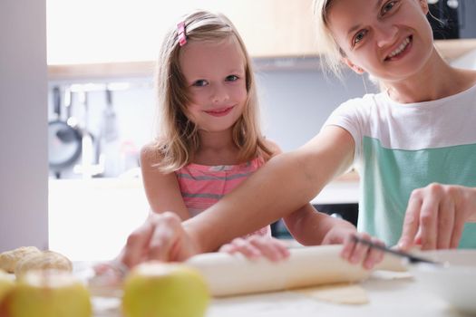 Happy young mother and little child daughter rolls out dough on kitchen table with pin. Cooking family meal for an adult and child and joyful emotions