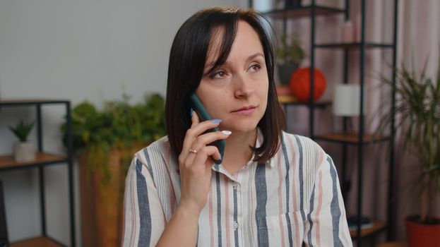 Young woman freelancer sits at desk at home office answering to client on mobile phone call, using smartphone. Conversation. Adult pretty girl student study working online remote distance job, talking