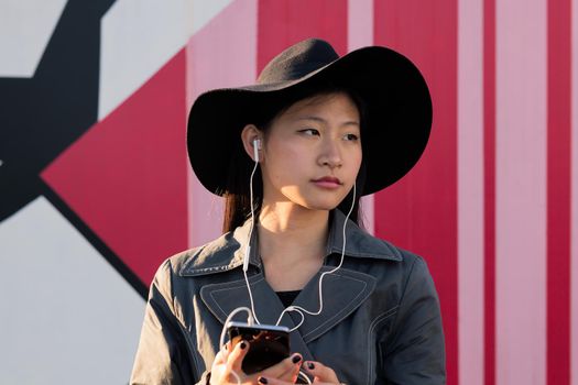 young beautiful classy asian woman with earphones listening music from her phone in a pink background, concept of technology and fashion lifestyle