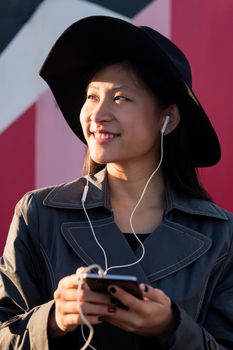 vertical portrait of a smiling beautiful classy asian woman with earphones listening music from her phone, concept of technology and fashion lifestyle