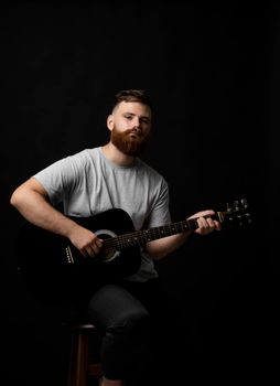 Bearded brutal guitarist plays an acoustic guitar in a black room and looking in the camera. The concept of music recording, rehearsal or live performance