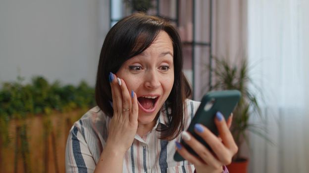 Wow. Happy winner woman freelancer use mobile smartphone typing browsing say Yes found out great big win good news celebrate victory sitting at home office. Girl student working online distance job